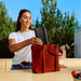 Black Sonos Play Portable Bluetooth Speaker being lowered into a red tote bag by a woman standing at an outdoor counter with trees behind her