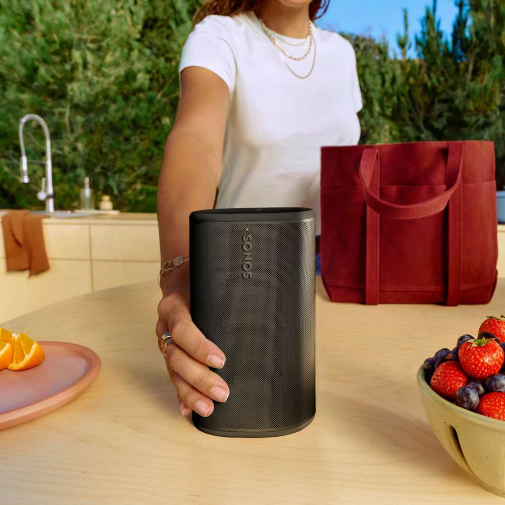 Black Sonos Play Portable Bluetooth Speaker held in one hand on an outdoor kitchen counter beside a bowl of strawberries and a red tote bag