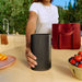Black Sonos Play Portable Bluetooth Speaker held in one hand on an outdoor kitchen counter beside a bowl of strawberries and a red tote bag
