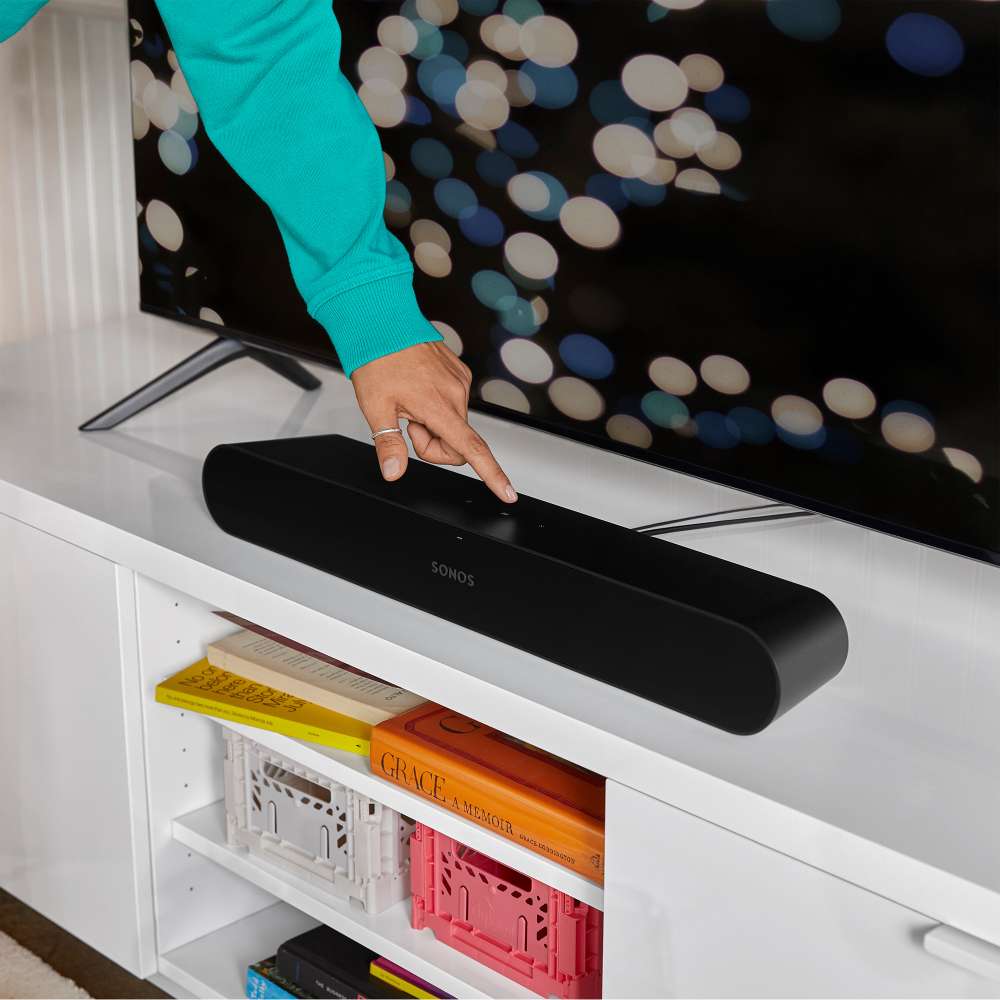 Black Sonos Ray Sound Bar a hand tapping the top of the bar as it sits on a white media unit below a television, with colourful storage crates on the shelf underneath