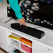 Black Sonos Ray Sound Bar a hand tapping the top of the bar as it sits on a white media unit below a television, with colourful storage crates on the shelf underneath