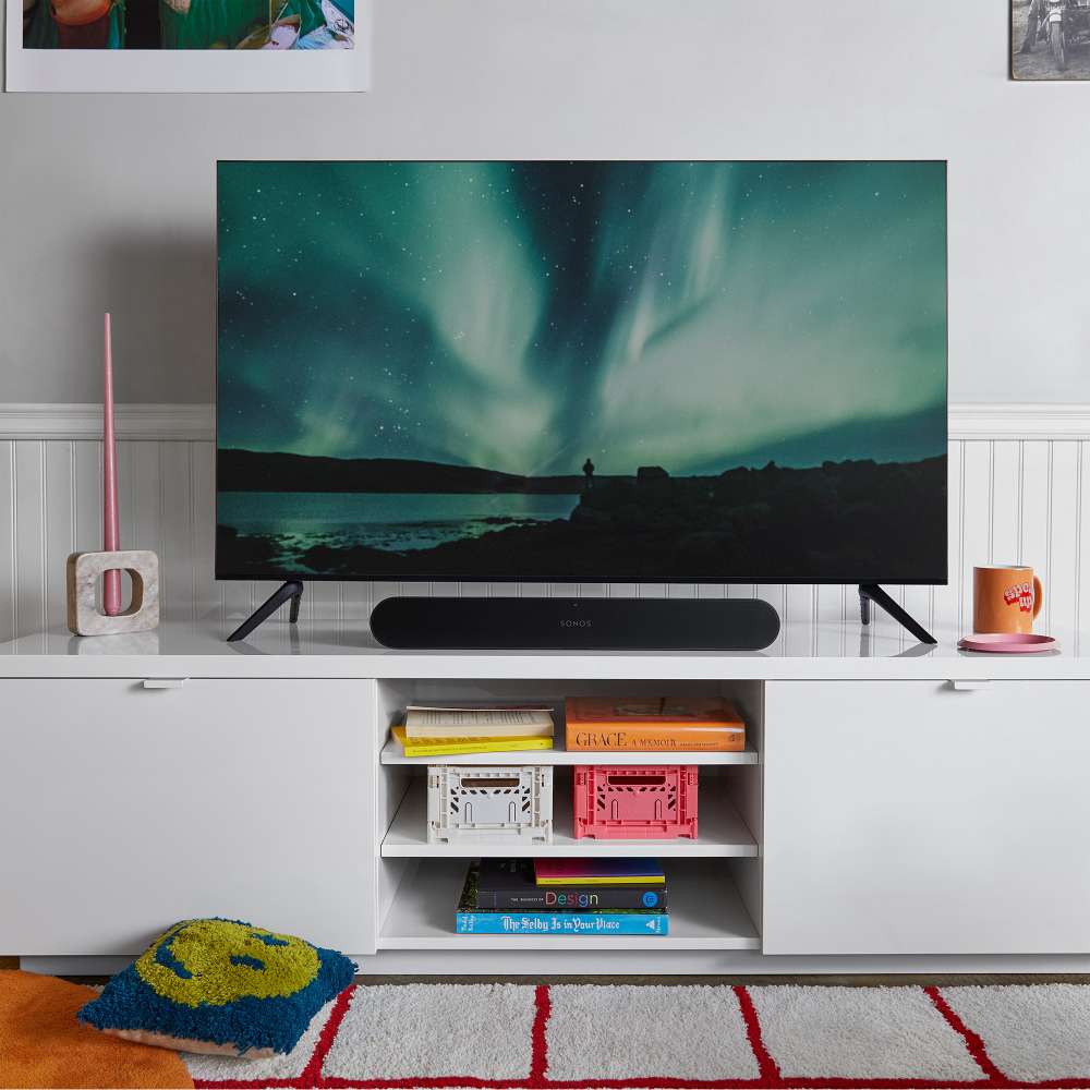 Black Sonos Ray Sound Bar sitting in front of a television showing the northern lights on a white sideboard in a living room with a striped rug
