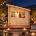 Centered front view of the illuminated Bluewater Den 67 on a clear Nevada night. Two adults watch sports inside while pathway lights, string lights, desert plants, a fire feature, and distant mountains surround the compact office.
