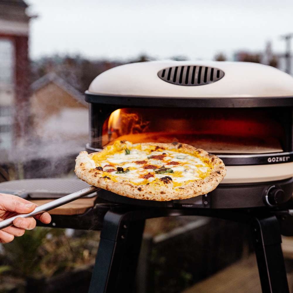 Bone Gozney Arc XL Pizza Oven outdoors on a black stand with a freshly baked pizza lifted out on a peel and steam rising in a garden setting.