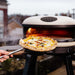 Bone Gozney Arc XL Pizza Oven outdoors on a black stand with a freshly baked pizza lifted out on a peel and steam rising in a garden setting.