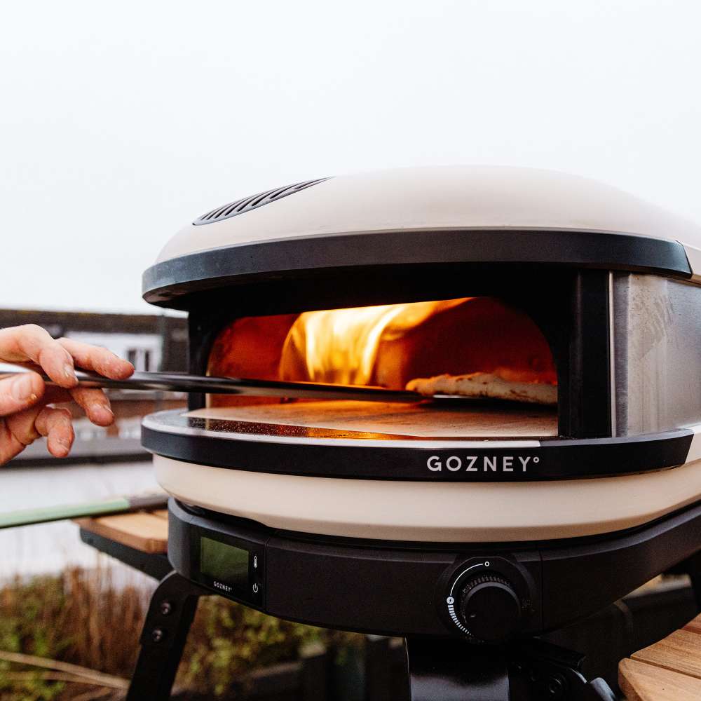 Bone Gozney Arc XL Pizza Oven outdoors on a black stand with two hands sliding a pizza into the flaming mouth on a long metal peel.