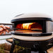 Bone Gozney Arc XL Pizza Oven outdoors on a black stand with two hands sliding a pizza into the flaming mouth on a long metal peel.