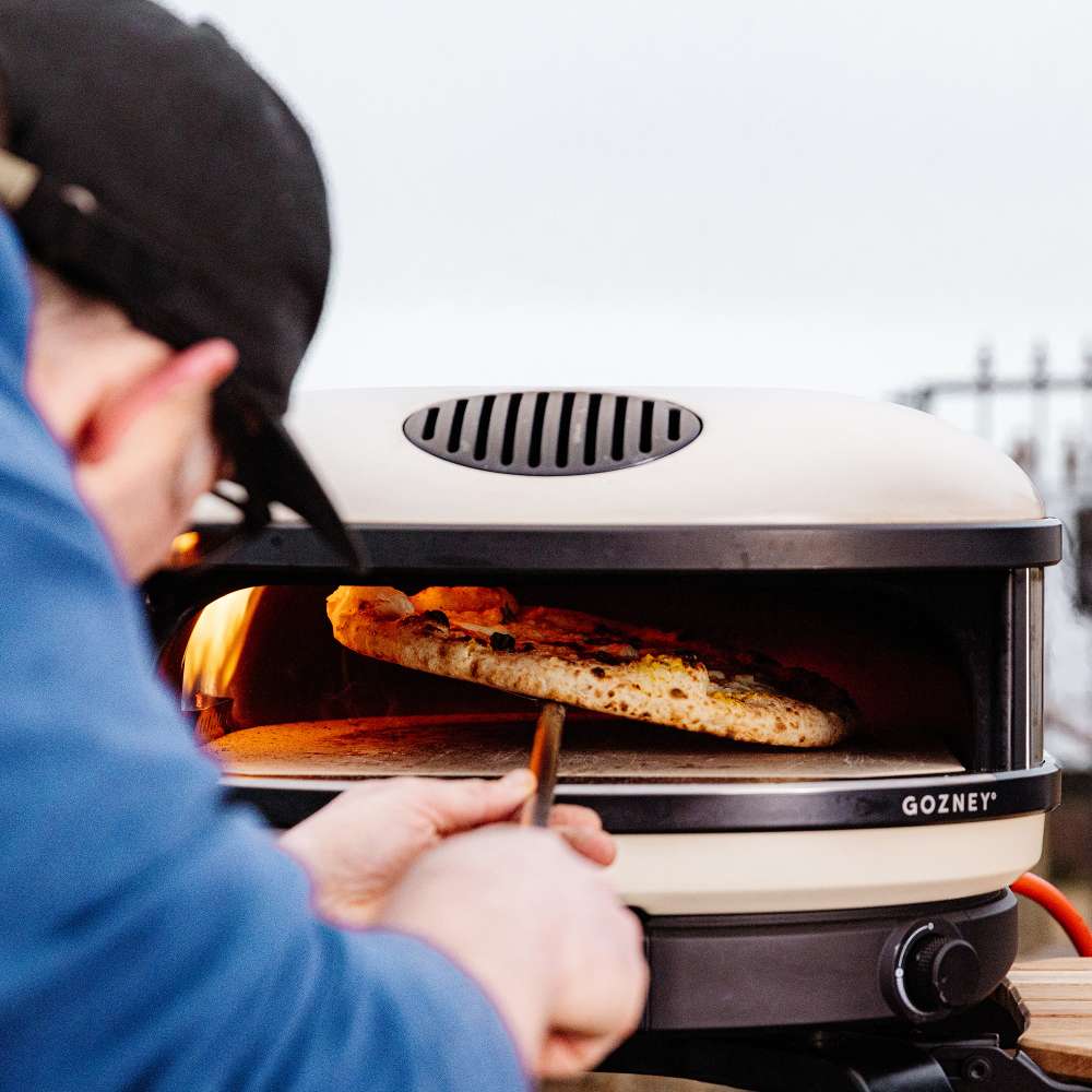 Bone Gozney Arc XL Pizza Oven with a person in a blue jacket and black cap sliding a pizza into the flaming mouth on a wooden peel.