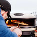 Bone Gozney Arc XL Pizza Oven with a person in a blue jacket and black cap sliding a pizza into the flaming mouth on a wooden peel.