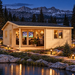 Light wood cabin at night with glowing windows, people inside, and snowy mountains in the background.
