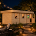 Rectangular light wood studio with flat roof, glass windows, and open door at night next to a stone path and fire pit.