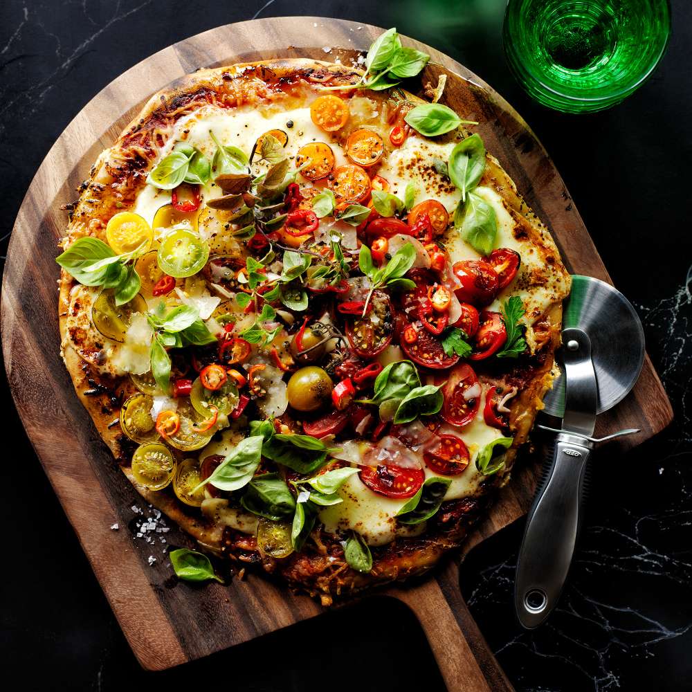 Crate & Barrel Acacia Pizza Peel on dark marble holding a pizza topped with heirloom tomatoes, mozzarella, red chilies and basil, with flaky salt scattered on the board, a stainless steel wheel cutter beside it and a green glass in the corner.