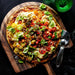 Crate & Barrel Acacia Pizza Peel on dark marble holding a pizza topped with heirloom tomatoes, mozzarella, red chilies and basil, with flaky salt scattered on the board, a stainless steel wheel cutter beside it and a green glass in the corner.
