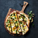 Crate & Barrel Acacia Pizza Peel on dark slate holding a sliced flatbread topped with squash, burrata, lemon and fresh herbs, the tapered handle and black leather cord at the upper right.