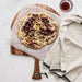 Crate & Barrel Acacia Pizza Peel seen from above on a pale plaster surface, carrying a flatbread on parchment topped with roasted grapes, figs, blue cheese, walnuts and honey, with a glass of red wine and a striped linen towel alongside.