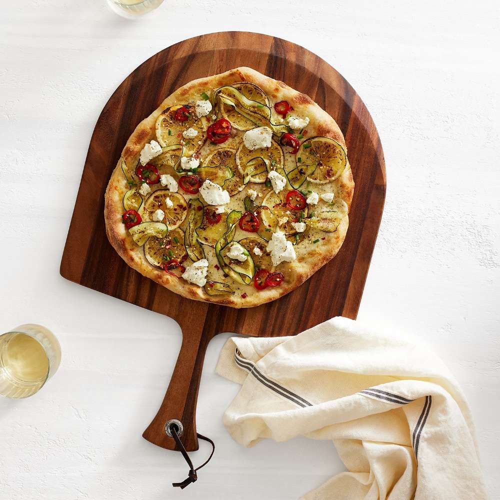 Crate & Barrel Acacia Pizza Peel seen from above on a white surface, holding a pizza topped with zucchini ribbons, lemon slices, ricotta and red chilies, with a glass of white wine on one side and a striped linen towel on the other.