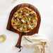Crate & Barrel Acacia Pizza Peel seen from above on a white surface, holding a pizza topped with zucchini ribbons, lemon slices, ricotta and red chilies, with a glass of white wine on one side and a striped linen towel on the other.