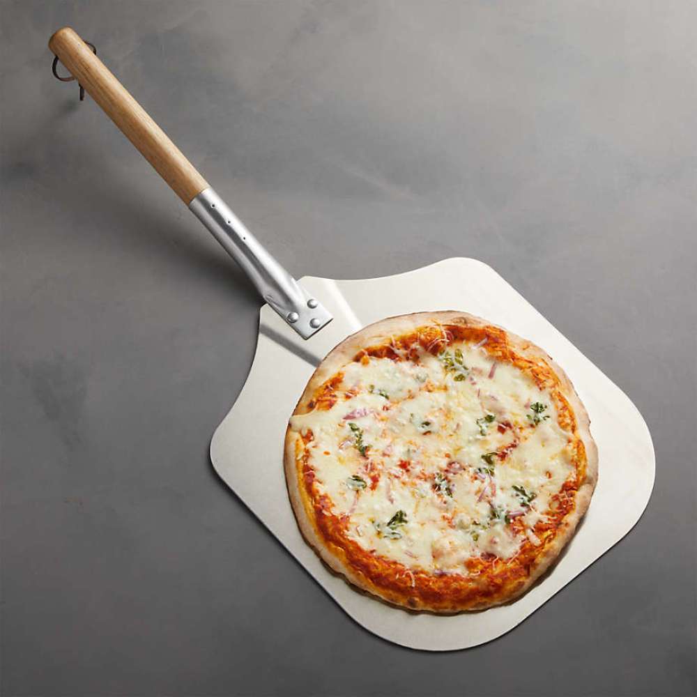 Crate & Barrel Metal Pizza Peel holding a whole cheese pizza on a gray countertop, seen from above with the wood handle and leather loop extending to the upper left.