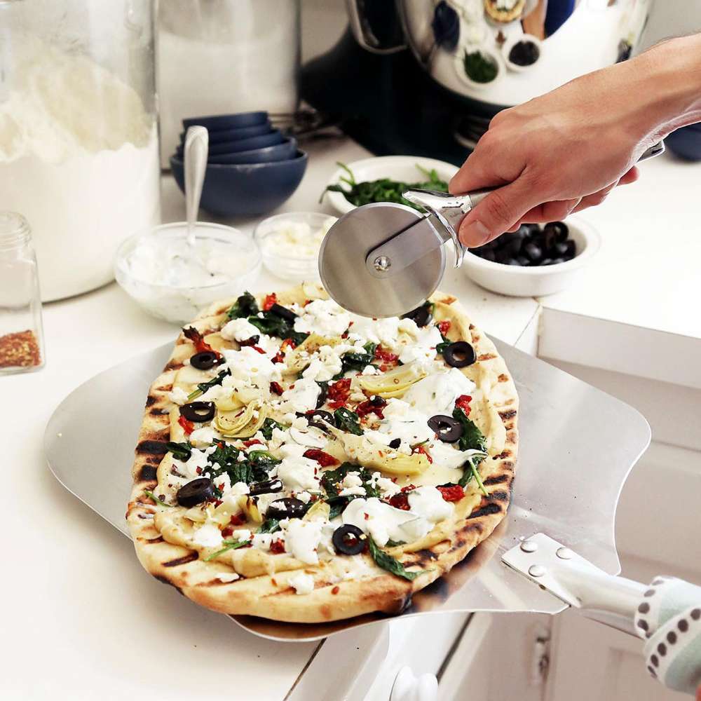 Crate & Barrel Metal Pizza Peel in a white kitchen carrying a grilled pizza topped with feta, black olives, artichokes and spinach, with a hand slicing another pizza with a wheel cutter and bowls of toppings behind.
