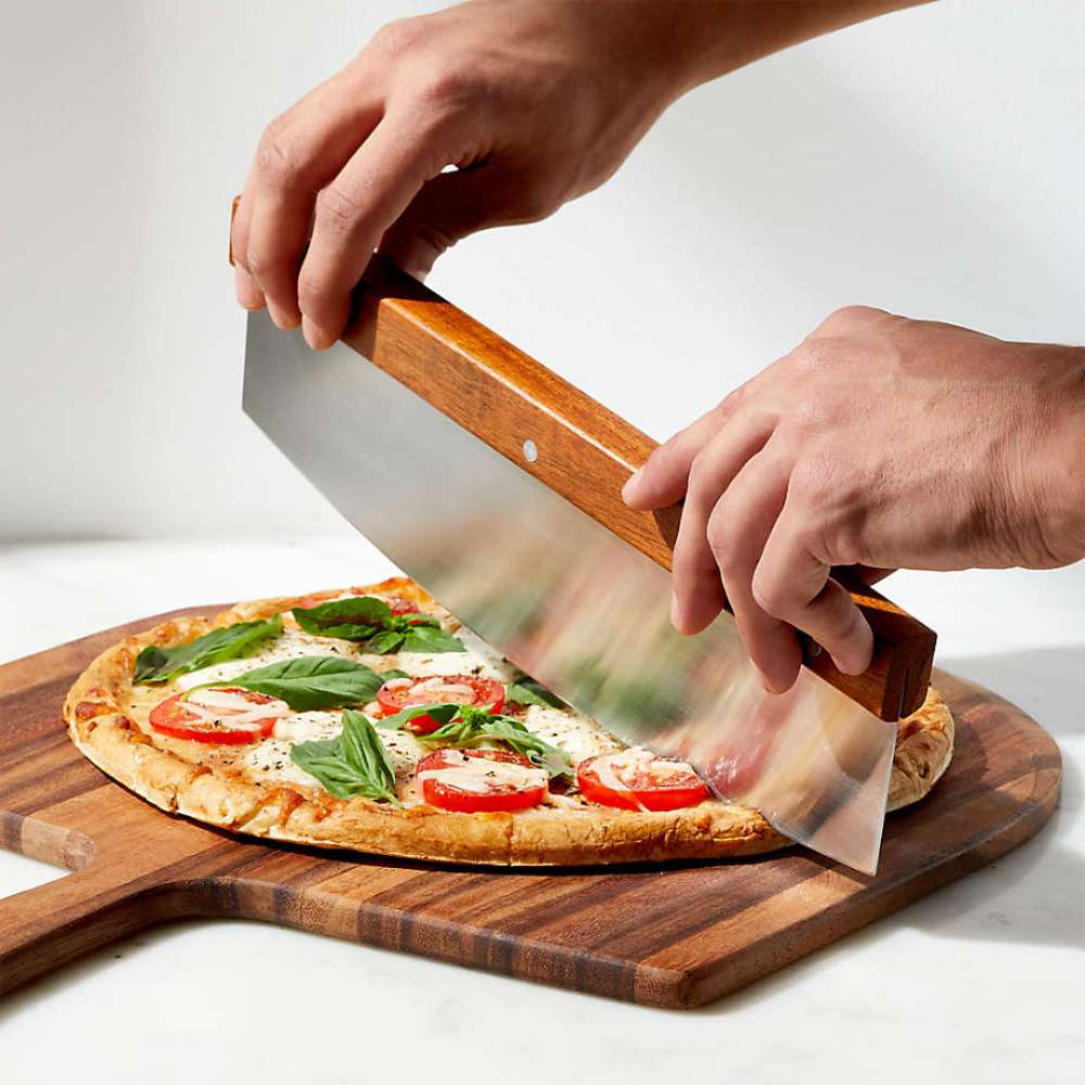 Crate & Barrel Rocker Pizza Cutter held in two hands and rocked through a margherita pizza with tomato, mozzarella and basil on an acacia wood board over a white marble counter.