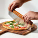 Crate & Barrel Rocker Pizza Cutter held in two hands and rocked through a margherita pizza with tomato, mozzarella and basil on an acacia wood board over a white marble counter.