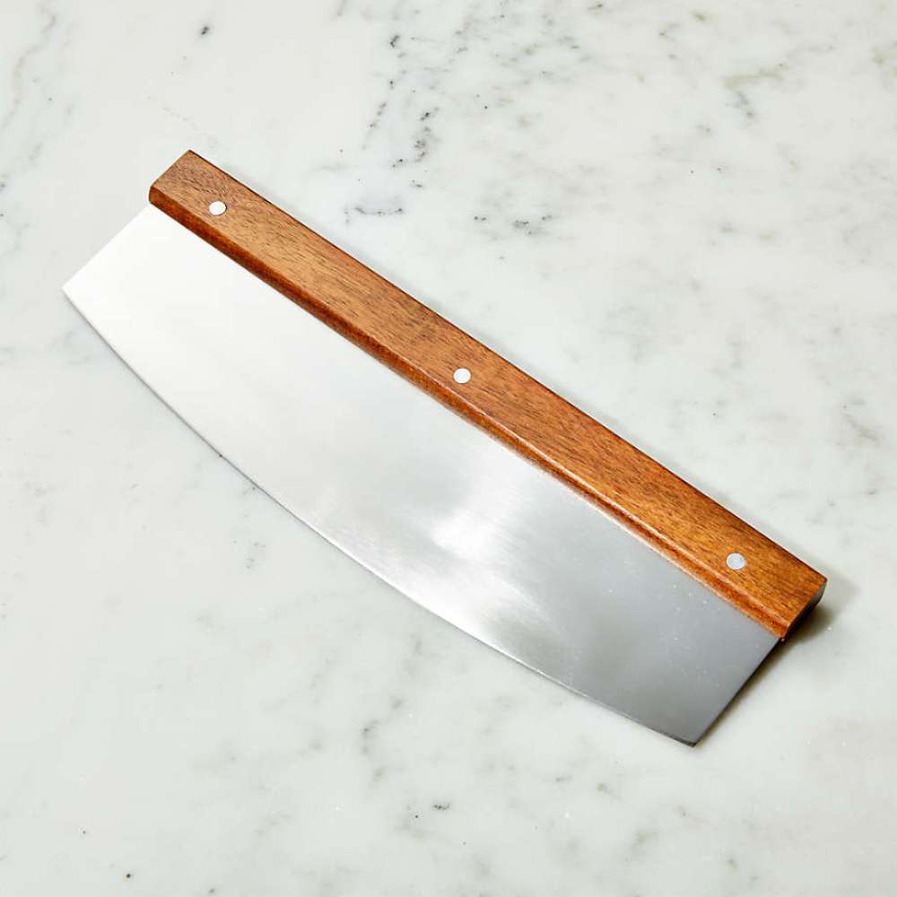 Crate & Barrel Rocker Pizza Cutter resting diagonally on a white marble countertop, the polished blade catching the light below the riveted wood handle.