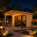 Small wood studio with glass doors and a porch, illuminated at night with people inside and a fire pit nearby.