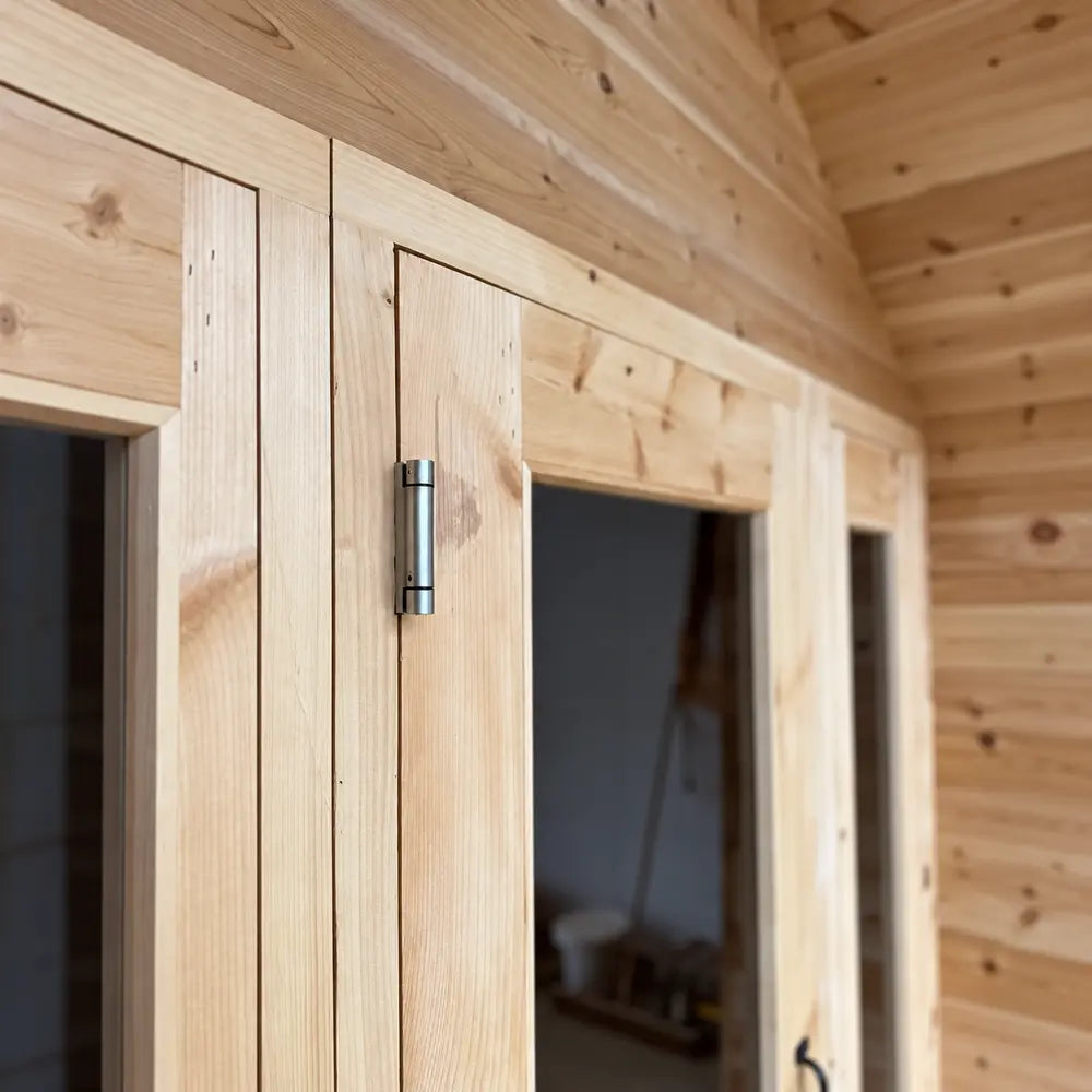 Close-up of a sturdy silver door hinge mounted on the smooth natural wood frame of the Sawmill Structures CabinSauna.