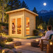 The warmly illuminated Versa Sauna sits beneath a bright moonlit Colorado sky, with two adults enjoying the sauna inside and another cooling down beside a firepit while facing the Rocky Mountains.