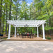 Centered white Structureworks Trex Pergola with built-in lighting, a ceiling fan, and a black dining set in a wooded patio setting.