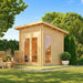 Sawmill Structures Versa Sauna in a sunny backyard with glass doors, a tall side window, wooden chairs, and lush landscaping.