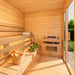 Bright TS TwoSauna interior with clean wood paneling, two-tier benches, under-bench lighting, sauna stones, a wooden bucket and ladle, folded towel, and natural garden light.
