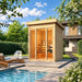 Sunny exterior view of the TS TwoSauna featuring sleek vertical wood construction, a full glass-front entrance, warm interior benches, and a landscaped poolside setting.