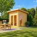 Sawmill Structures Versa Sauna glowing in warm sunlight beside a wooden dining table and chairs in a landscaped backyard.