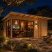 Rectangular wood studio with glass walls at night, showing people watching a game inside on a stone patio.