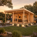 Rectangular wood studio with a flat roof, glass doors, and a lit interior bar set in a garden at sunset.