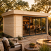 Light wood rectangular studio with glass sliding doors, set on a stone patio with a fire pit and trees at sunset.