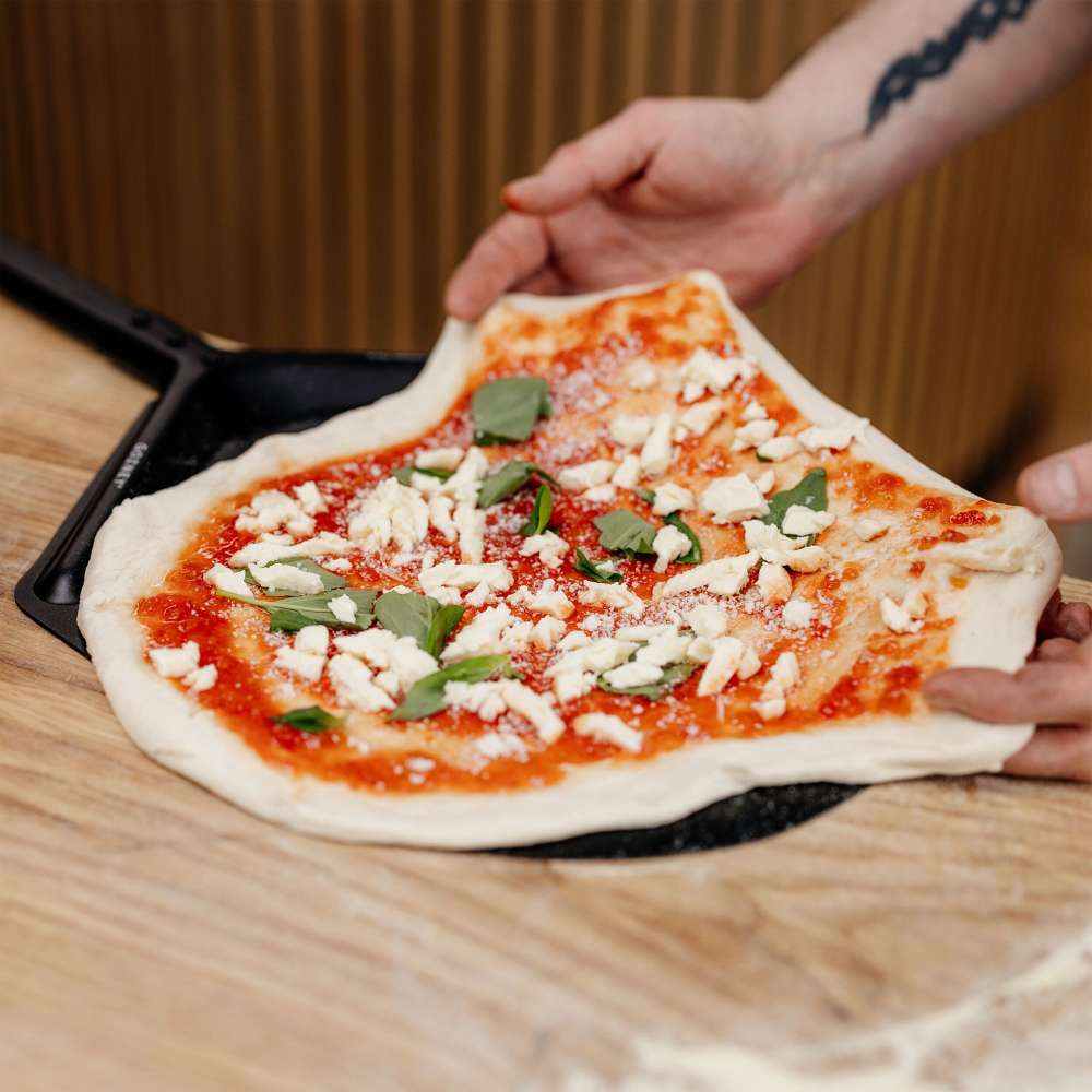 Gozney 14" Balance Placement Pizza Peel on a wooden counter with two hands easing a raw topped pizza onto the black perforated head.