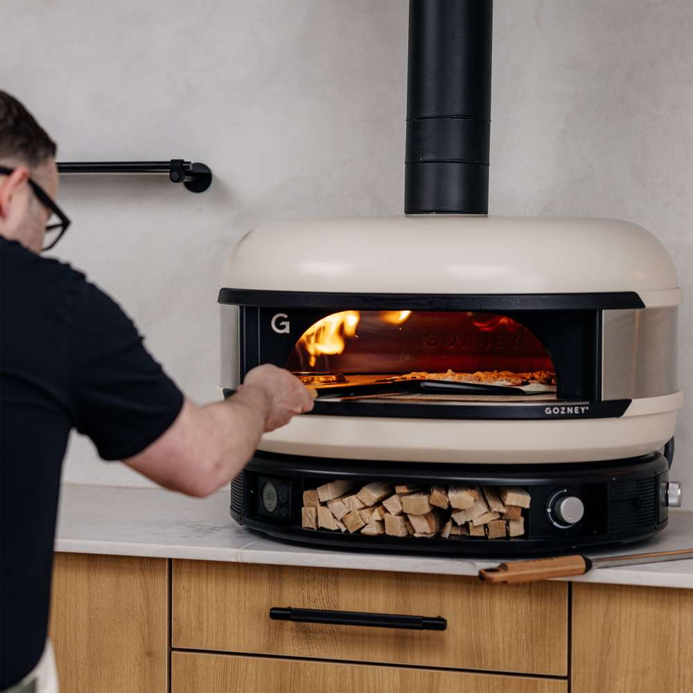 Gozney 14" Balance Placement Pizza Peel used by a man in a black tee to launch a pizza into a Bone Dome oven set in a counter with firewood stacked below.