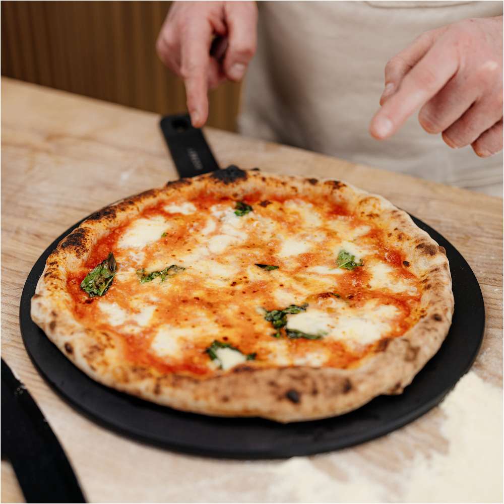 Gozney 14" Pizza Server held by a person in an apron on a countertop with a baked margherita pizza resting on it.