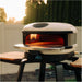 Gozney Arc XL Pizza Oven Mantel fitted to the mouth of a Bone Arc XL oven on a stand outdoors at sunset with a white fence and hedge behind.