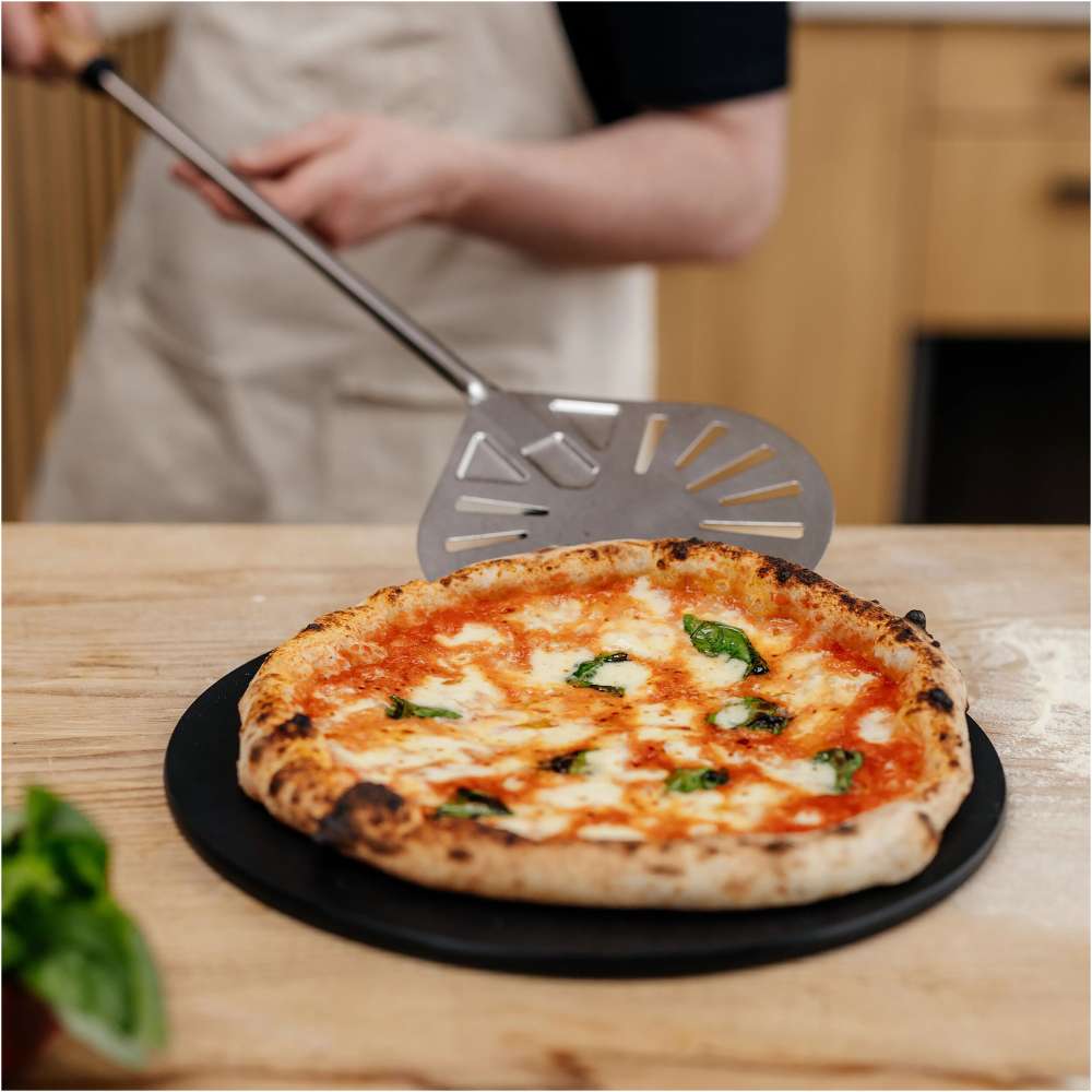 Gozney Balance Turning Peel held by a person in an apron above a finished margherita pizza on a black pan on a kitchen counter.
