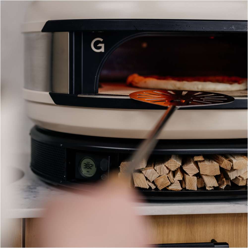 Gozney Balance Turning Peel turning a part-baked pizza inside a Bone Arc oven with firewood stacked on the shelf below.