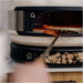 Gozney Balance Turning Peel turning a part-baked pizza inside a Bone Arc oven with firewood stacked on the shelf below.