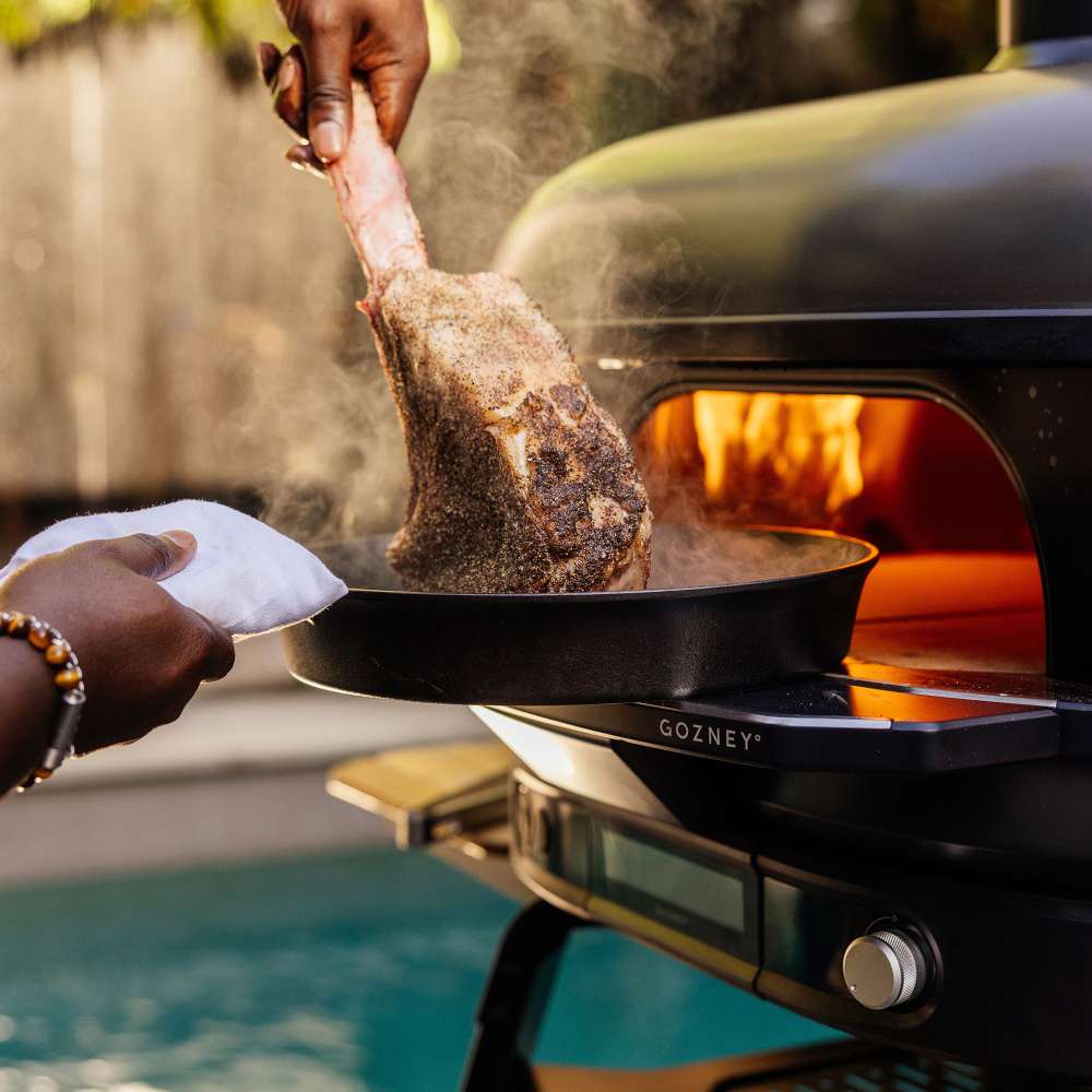 Gozney Dome 2 Mantel holding a cast iron pan as a hand lifts a seared tomahawk steak out of it at the glowing mouth of a Dome oven by a pool.