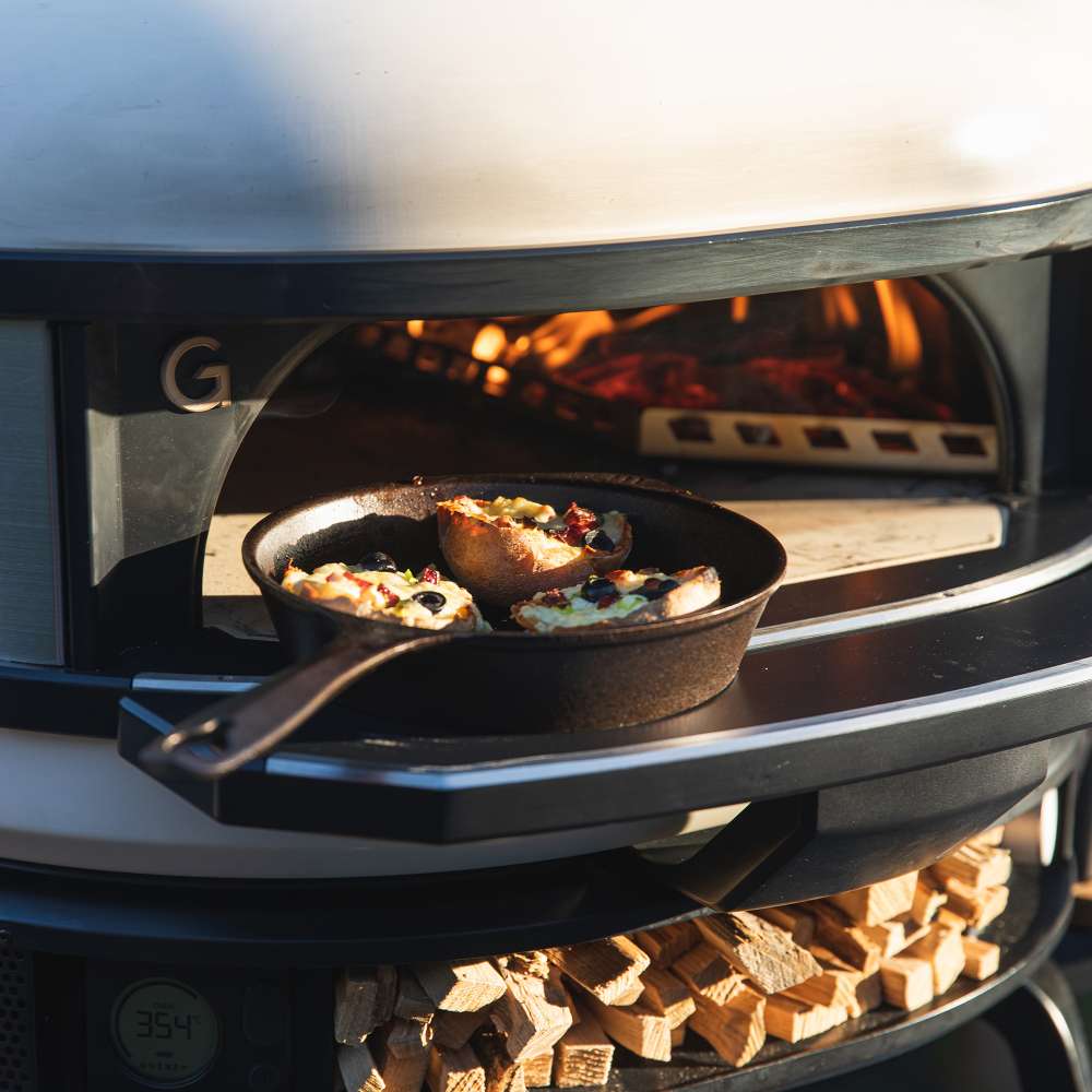 Gozney Dome 2 Mantel holding a cast iron skillet of food at the mouth of a Bone Dome oven with flames inside and firewood stacked below.