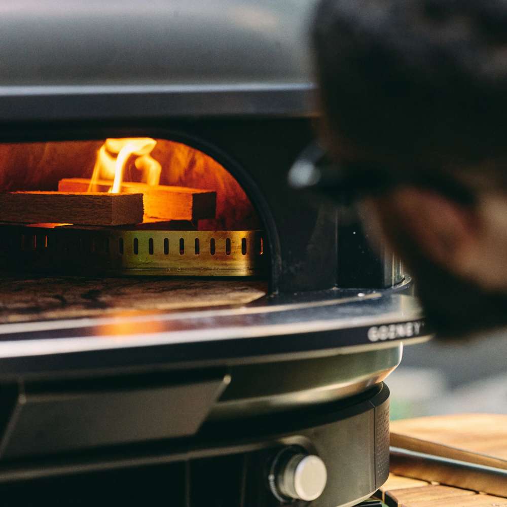 Gozney Dome Gen 2 Natural Gas Pizza Oven close view through the mouth with split logs burning on a stainless grate inside the glowing terracotta dome and a blurred person at the right.