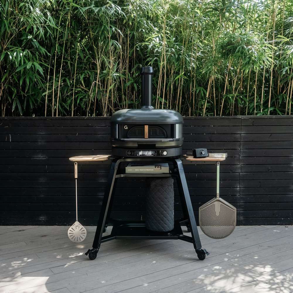Gozney Dome Gen 2 Natural Gas Pizza Oven on its black wheeled stand on a patio in front of a black slatted fence with bamboo behind and two peels hanging from the side hooks.