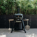 Gozney Dome Gen 2 Natural Gas Pizza Oven on its black wheeled stand on a patio in front of a black slatted fence with bamboo behind and two peels hanging from the side hooks.