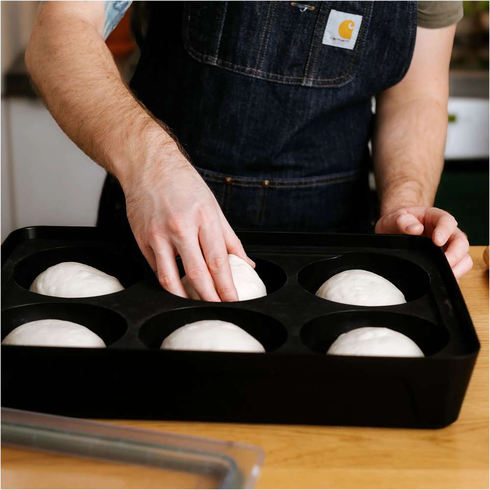 Gozney Pizza Dough Tray on a wooden countertop with a person in a denim apron setting a ball of dough into one of the six wells.